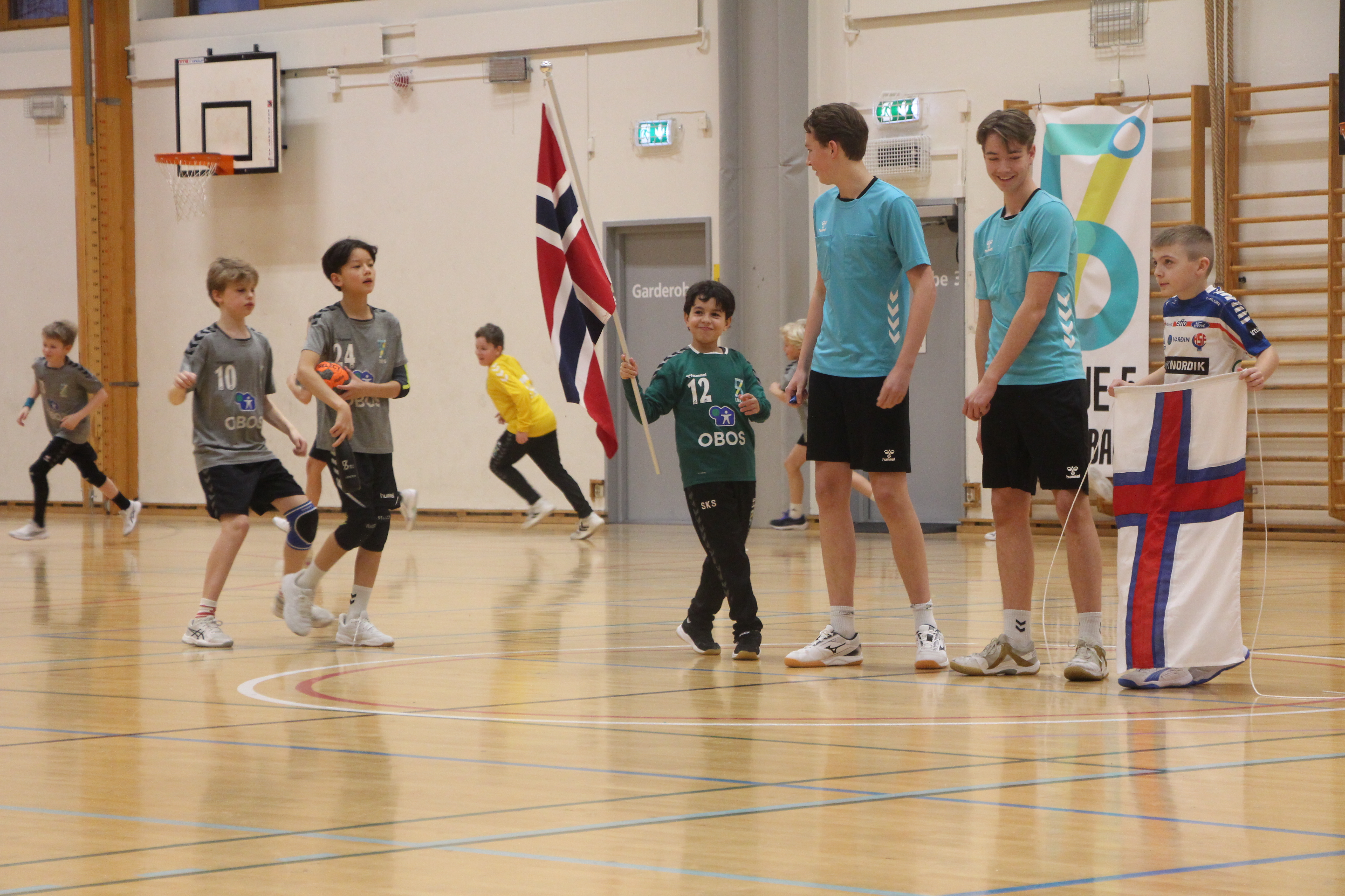 MINI-EM: Spillere fra Linje 5 og H71 samlet på banen i Linderudhallen før avkast, klare for uoffisiell landskamp mellom Norge og Færøyene med flagg, innmarsj og solid støtte fra tribunen.