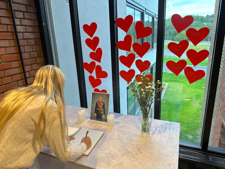 SISTE HILSEN: Annabelle skriver i skolens minneprotokoll. Rundt bildet av kong Harald har elevene hengt opp hjerter med personlige ord om kongen de har mistet.