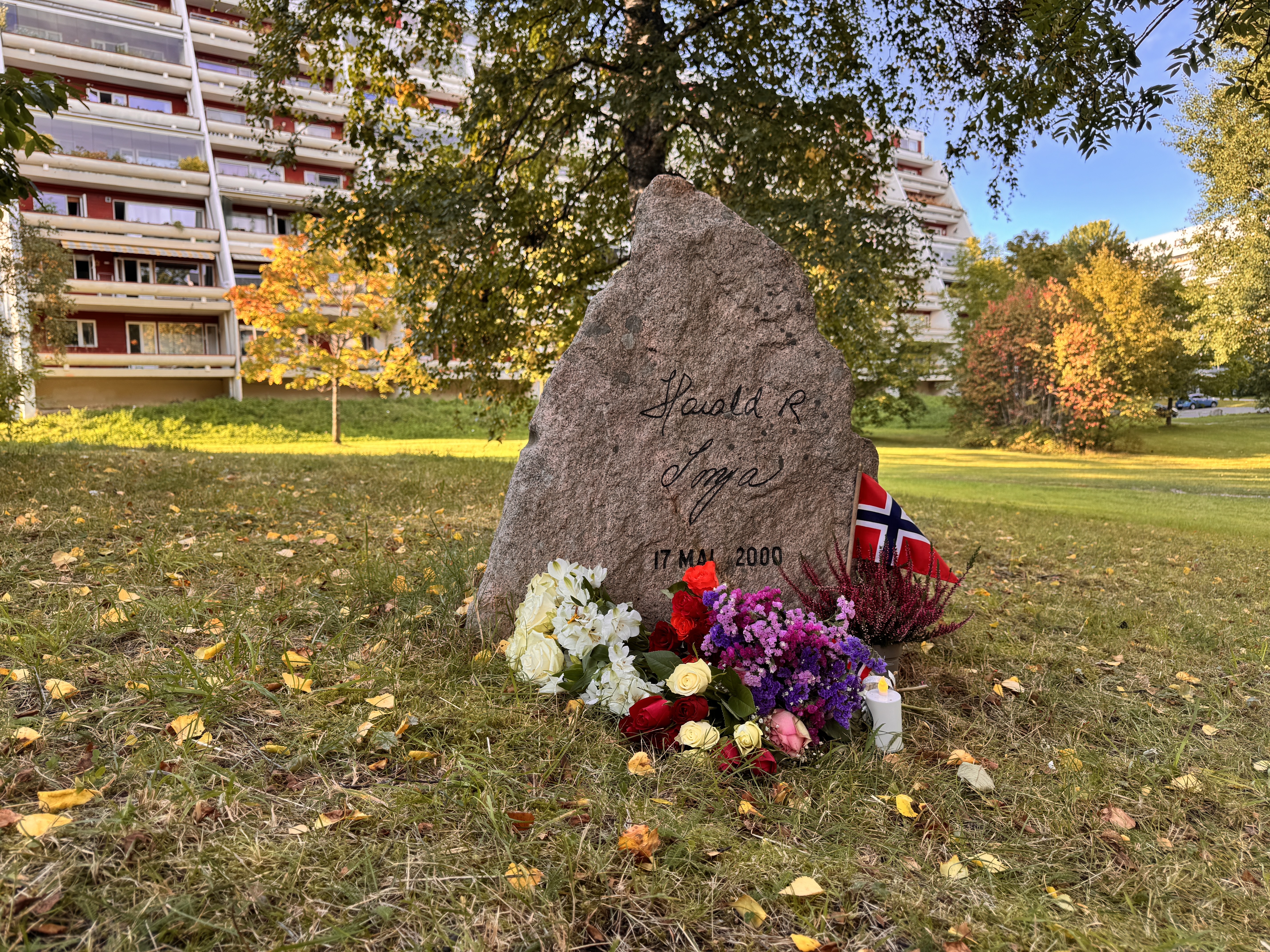 MINNES KONGEN: Blomster, norske flagg og lys er lagt ned ved steinen med signaturene til kong Harald og dronning Sonja. Steinen står ved tusenårstreet på kollen bak Fossum kirke og minner om kongeparets besøk på Stovner 17. mai 2000.